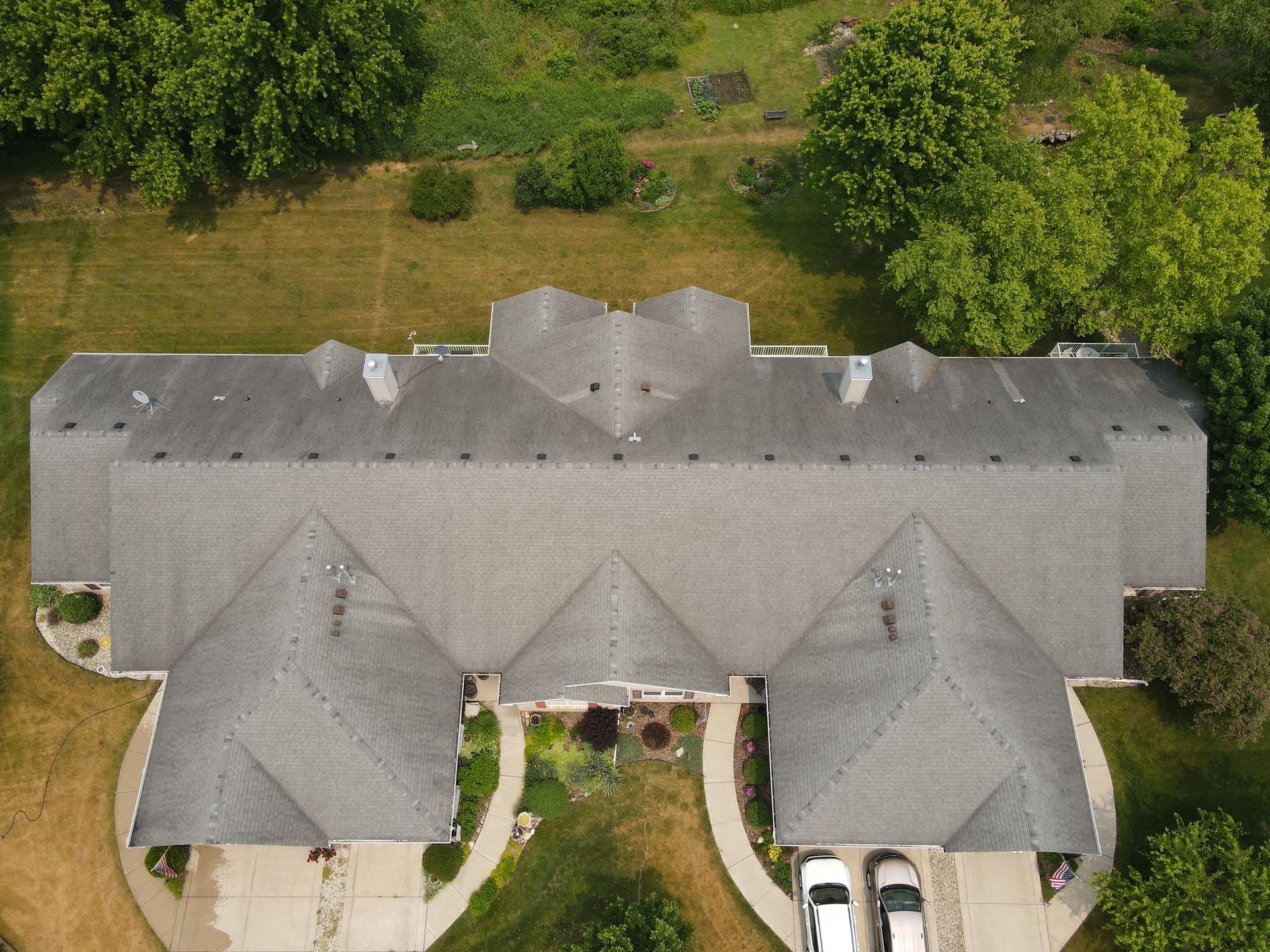 Overhead view of a long gray roofed house with cars parked in front, surrounded by trees and grass.