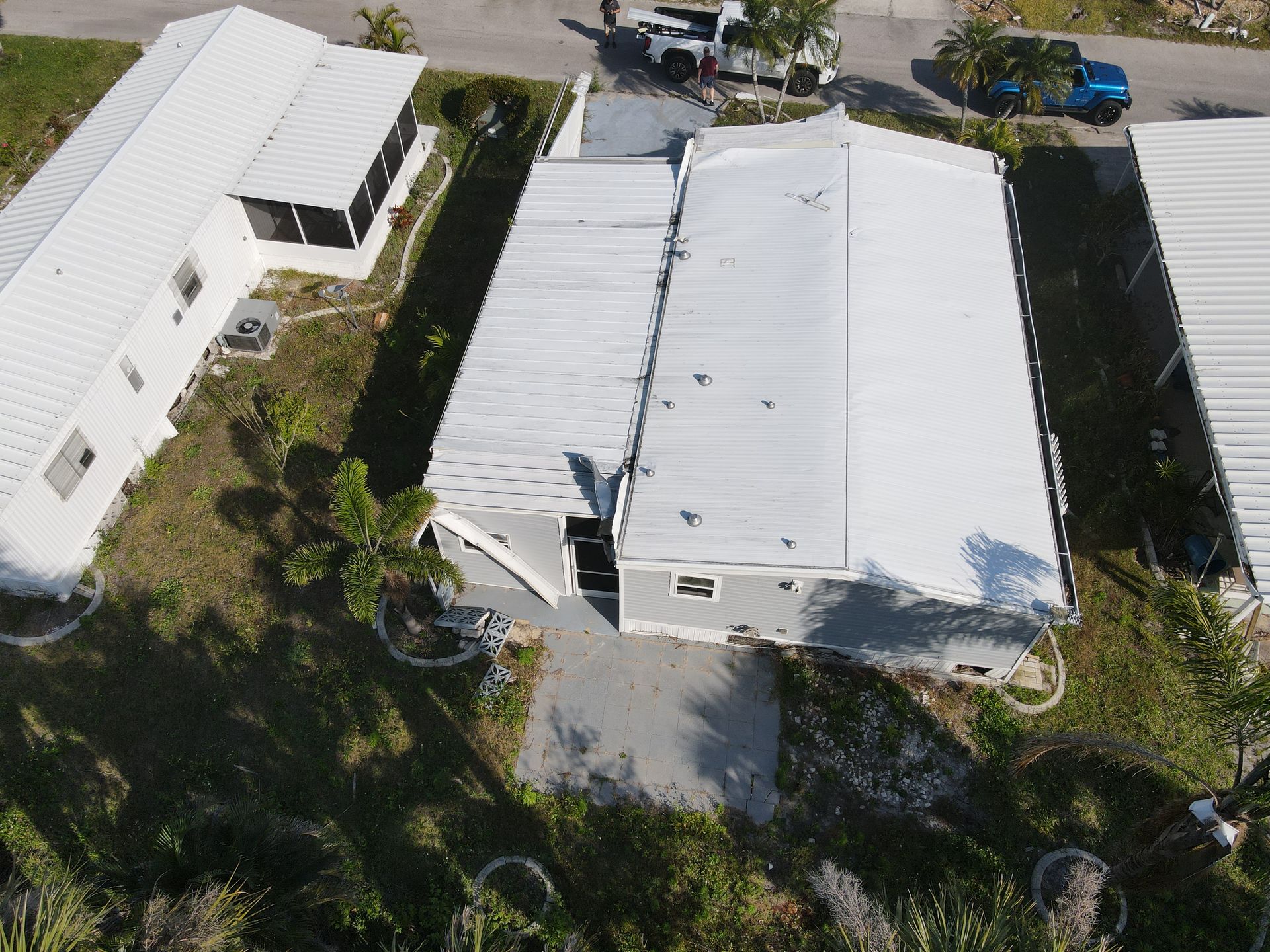 Aerial view of a gray house with a white roof surrounded by smaller white buildings and sparse green vegetation.