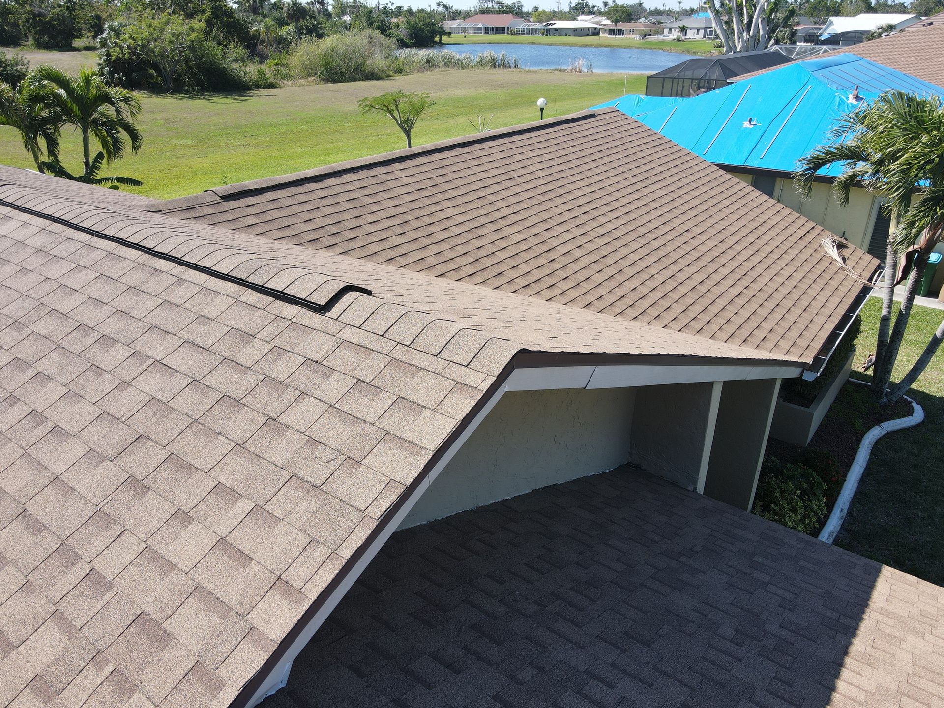 Brown shingled roof with a dark edge. Beige eaves are visible, green grass, trees, and a blue roof in background.