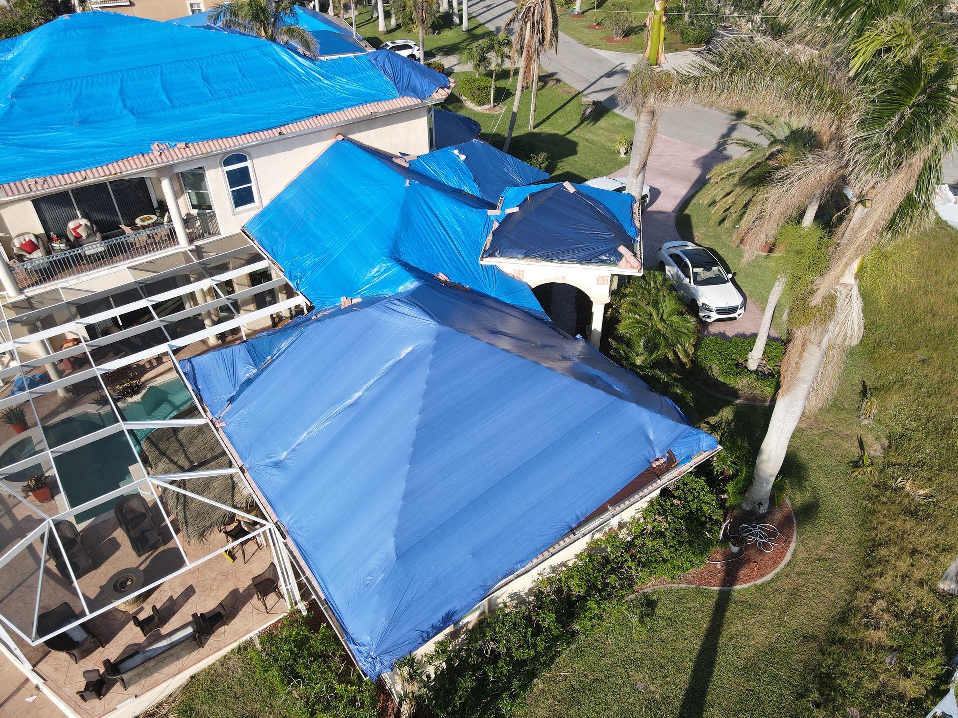 Blue tarp covers damaged roofs of a house after a storm; car in the driveway, palm tree in the yard.
