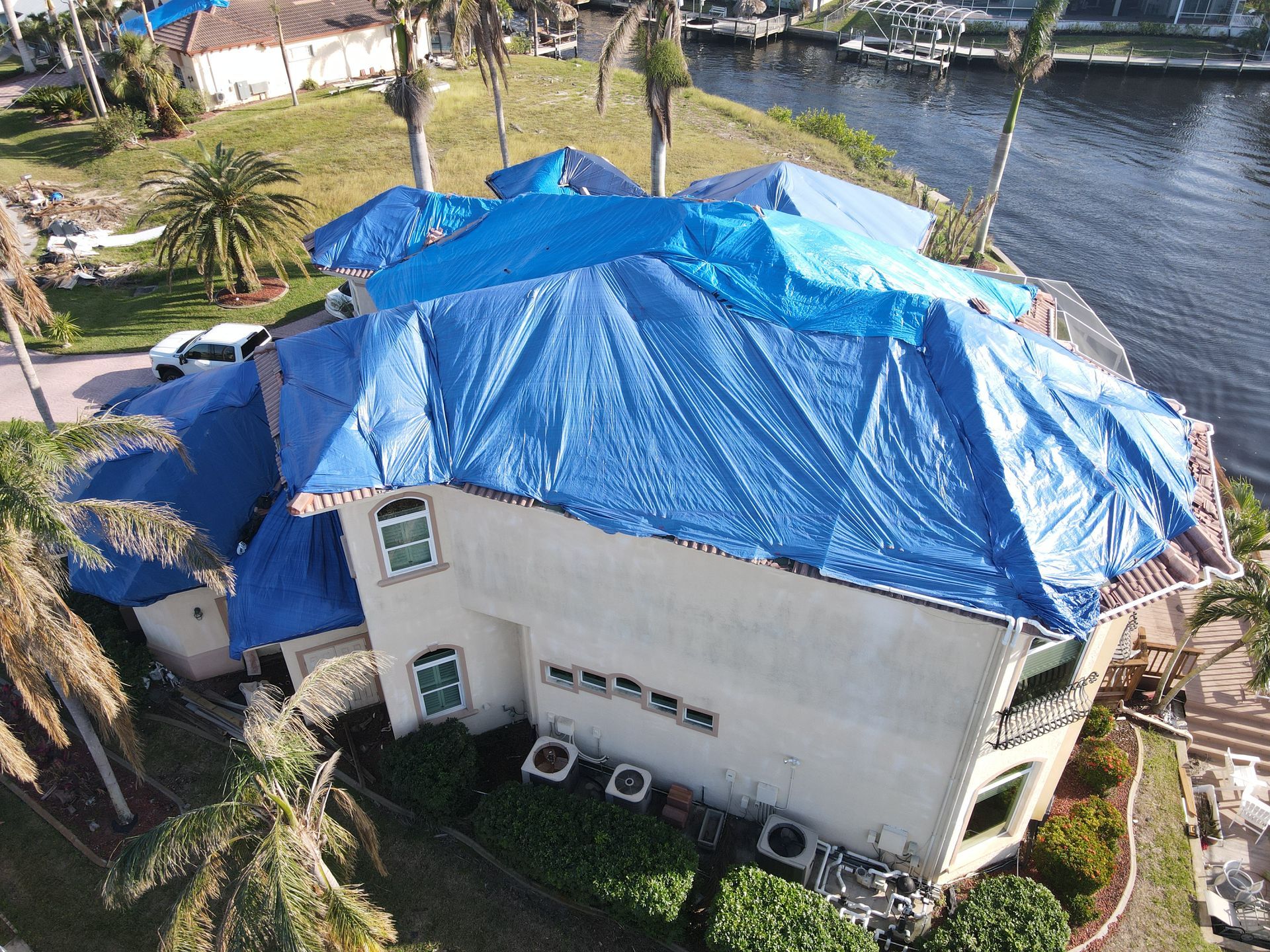 House with blue tarps covering the roof, likely damaged. Exterior shot near water, with palm trees.