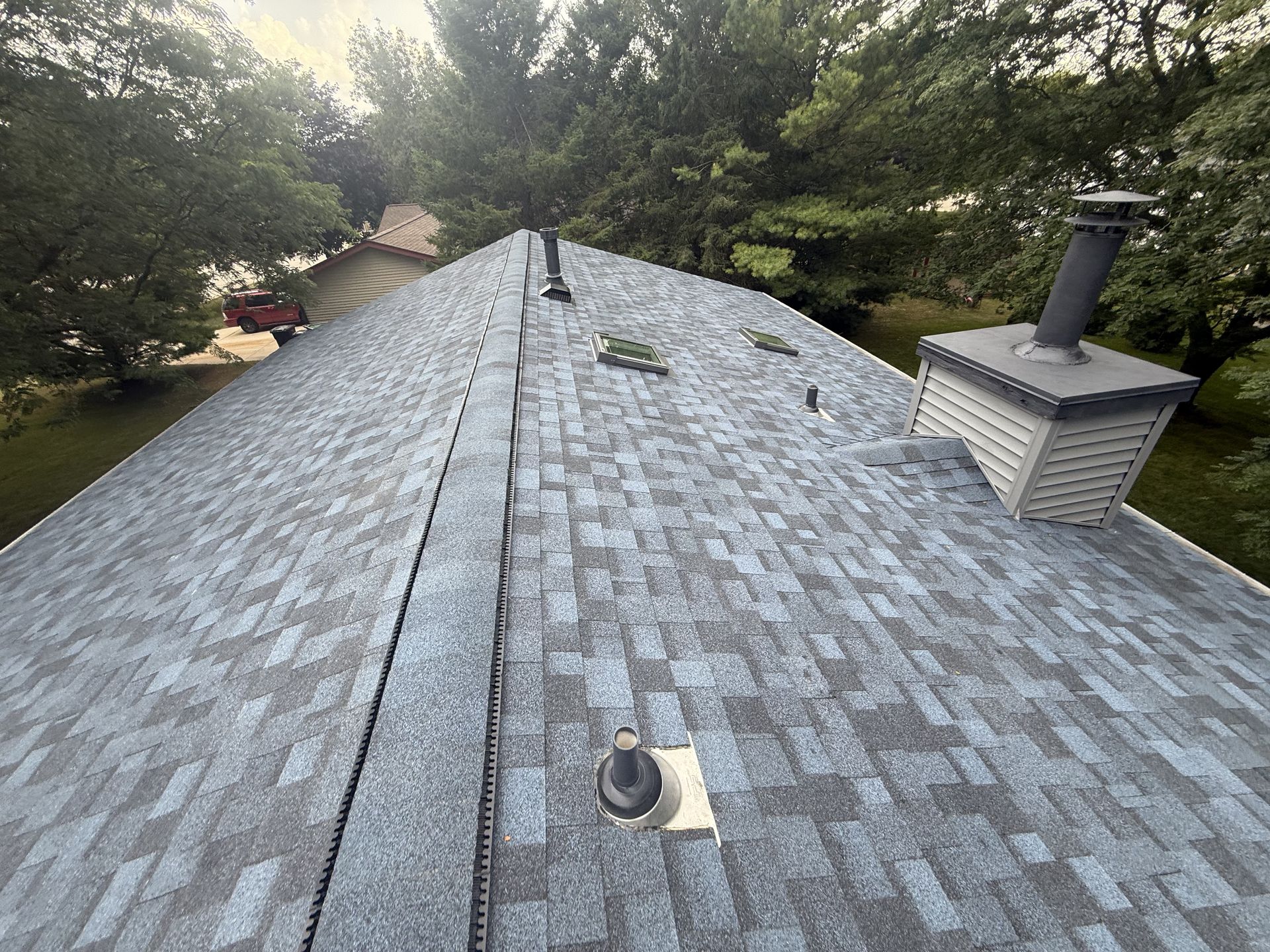 Overhead view of a gray shingle roof with a chimney and vents. Trees and sky in the background.