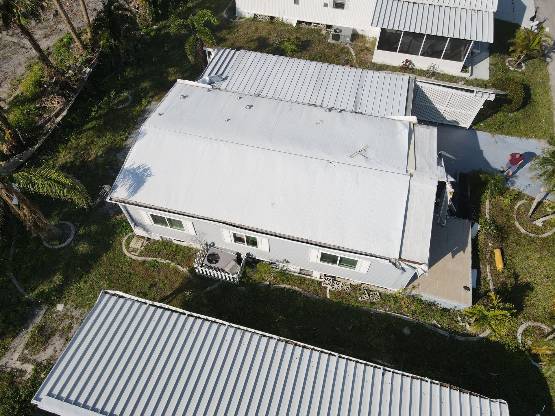Aerial view of a gray house with a metal roof surrounded by green grass and palm trees in a sunny setting.