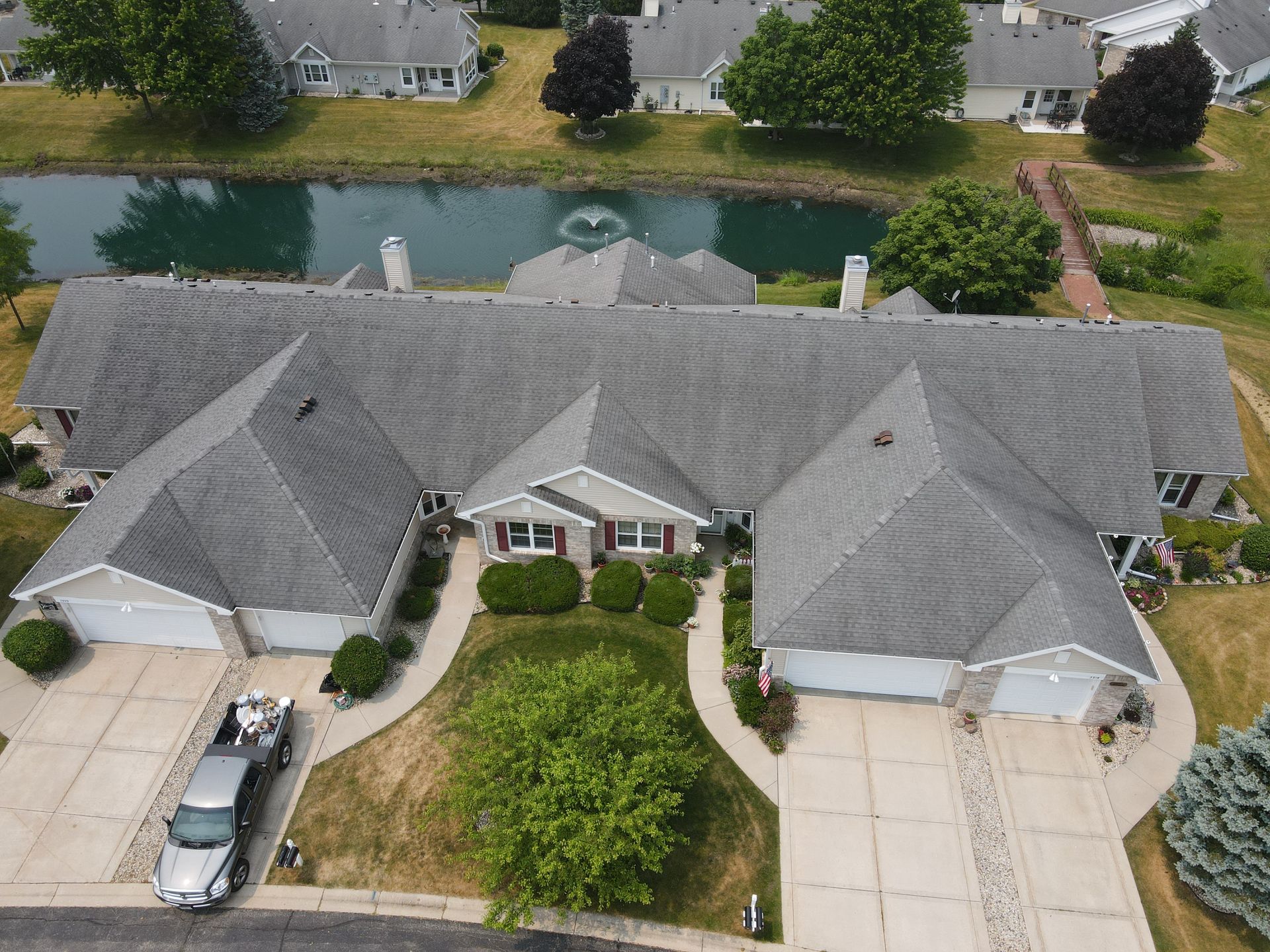 Aerial view of a gray-roofed townhouse complex with driveways, green lawns, and a pond in the background.