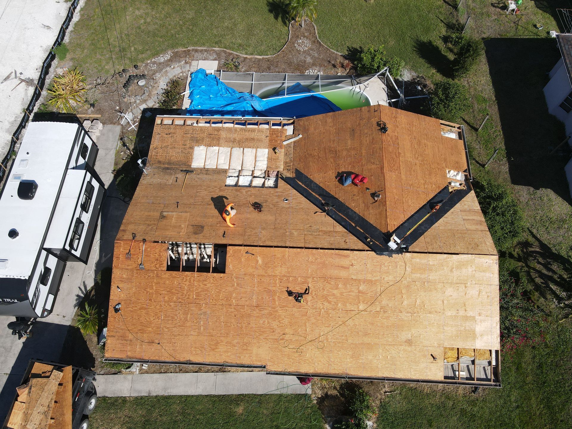 Aerial view of a house roof partially under construction. Workers on roof, blue pool visible in the background.