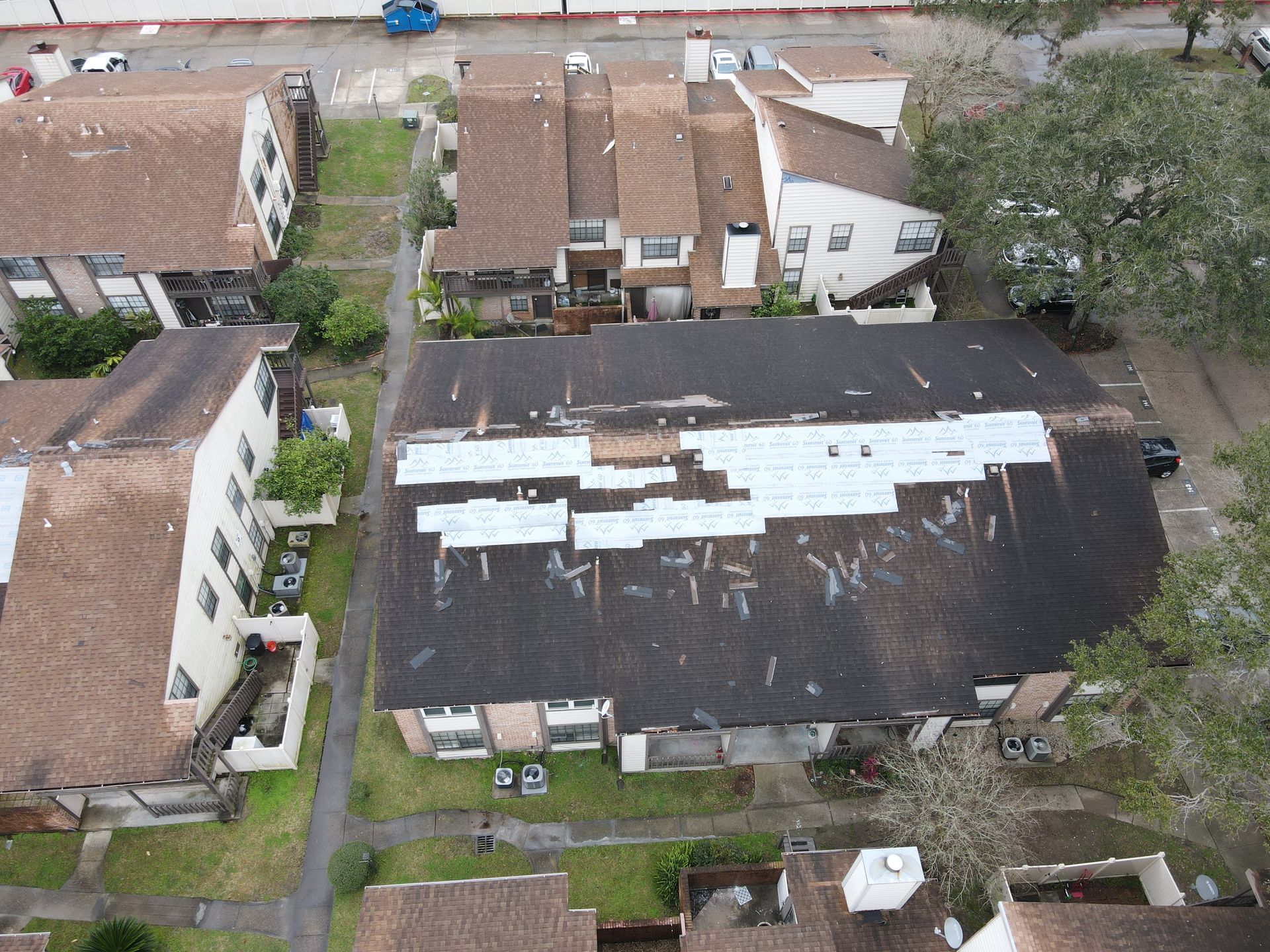 Overhead view of a row of townhomes with dark roofs. Patches of repair visible on one roof.