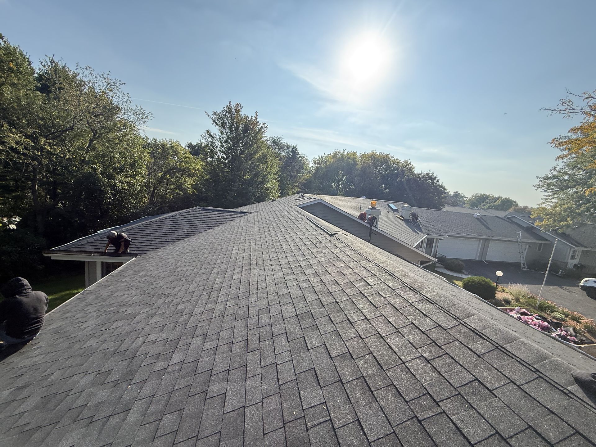 Roofers working on a shingled roof under a bright sunny sky with trees in the background.