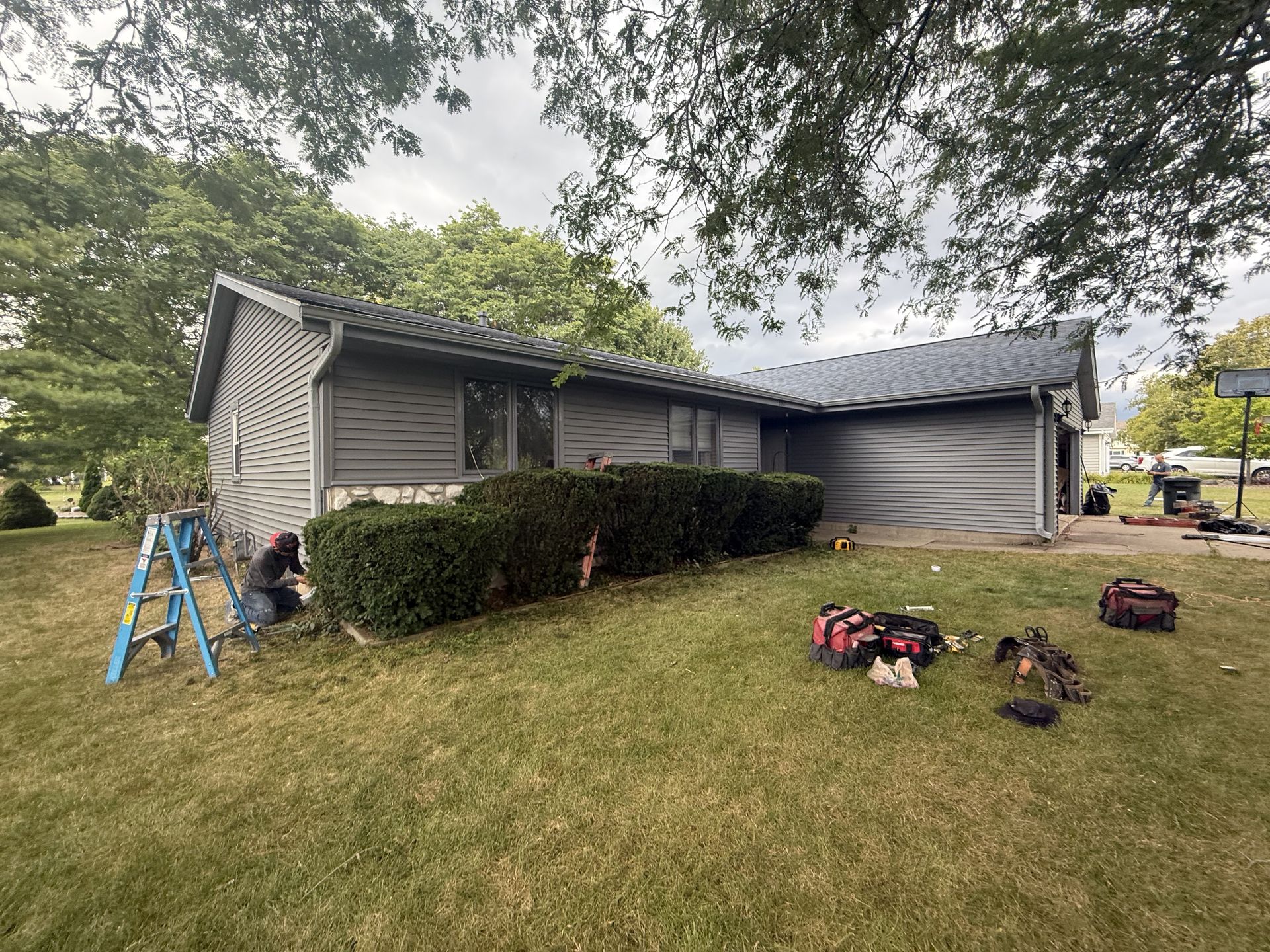 House with gray siding and roof, green bushes, and a worker on a ladder, overcast day.