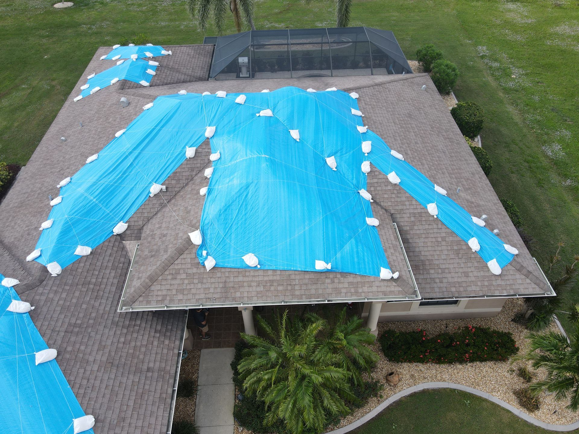 Blue tarps cover damaged roof of a house, secured with white tape. Aerial view.
