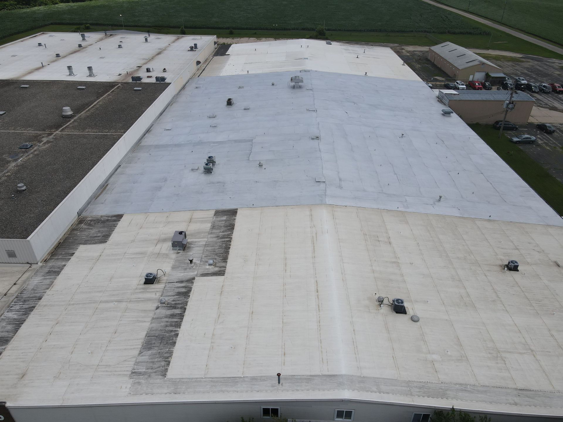 Aerial view of a large industrial building roof with varied white and gray sections, vents, and surrounding vegetation.