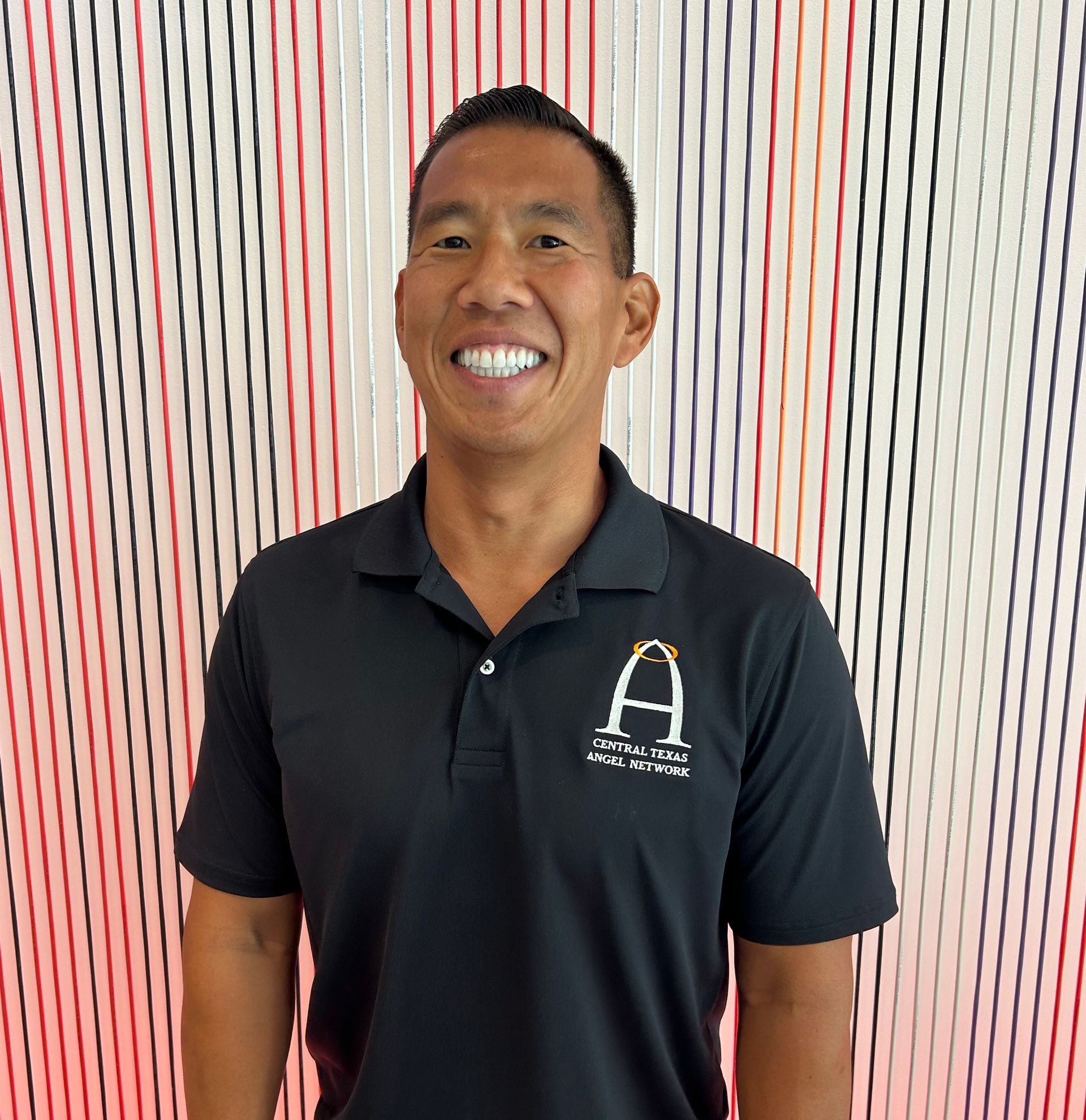 Smiling man in a black polo shirt with a logo, standing before a striped backdrop
