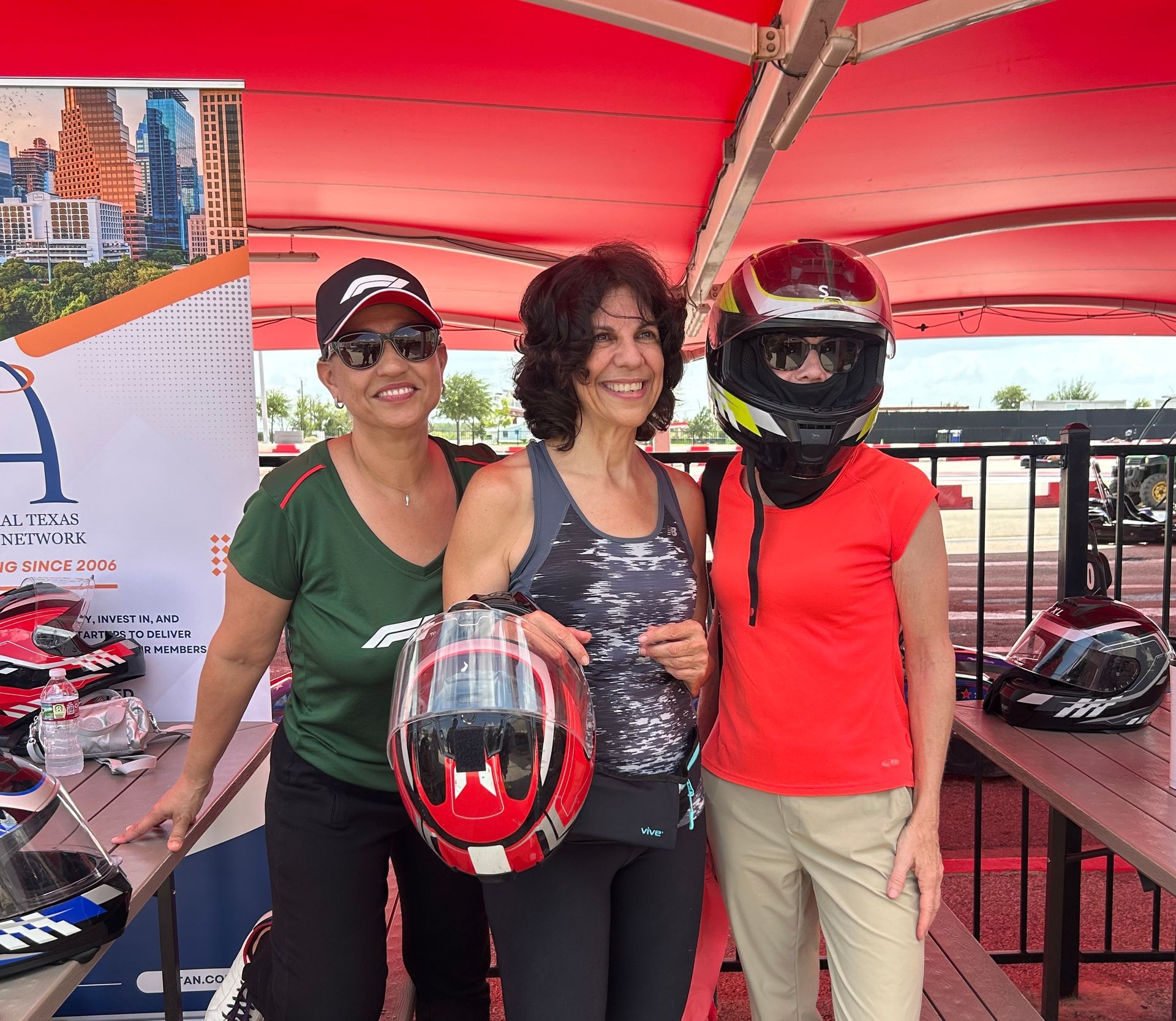 Three people at an outdoor go-kart track, smiling and holding helmets under a red canopy.