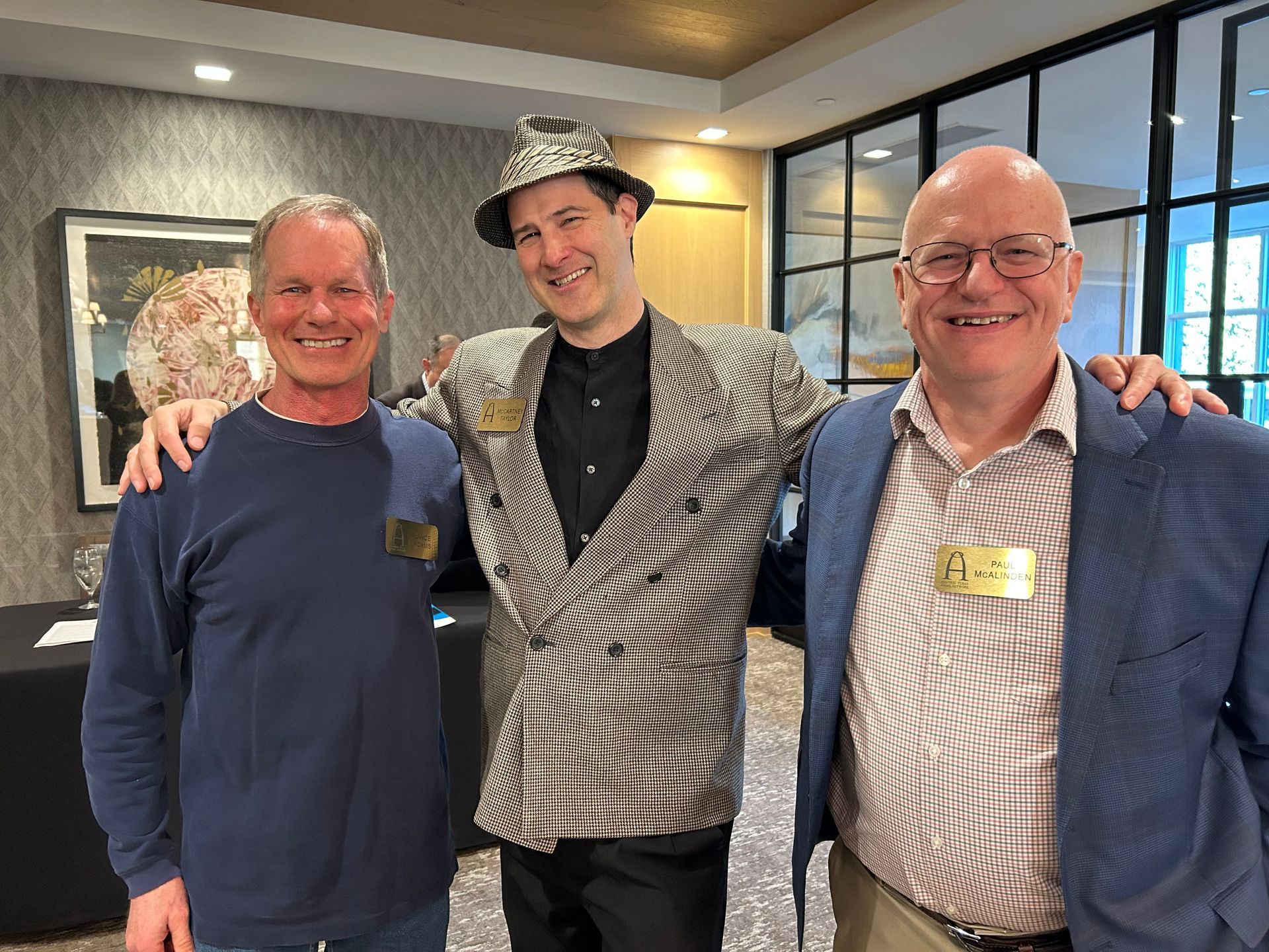Three smiling people pose together indoors at a reception, wearing business-casual clothes.