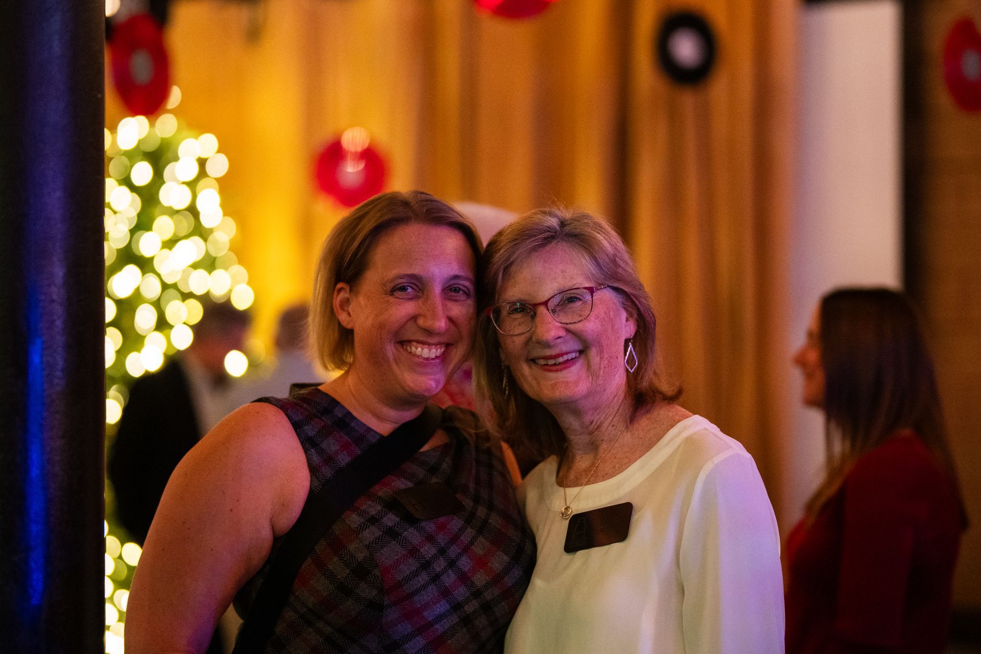 Two smiling women posing at a warmly lit indoor event with bokeh lights in the background