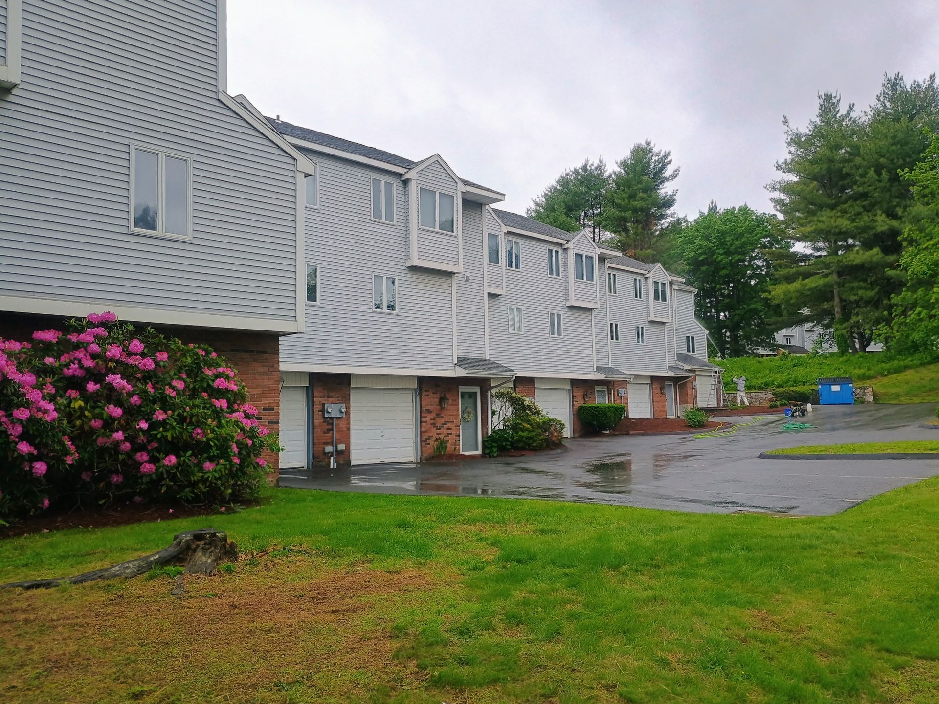 Row of light grey townhouses with attached garages, green lawn, and pink flowering bush.