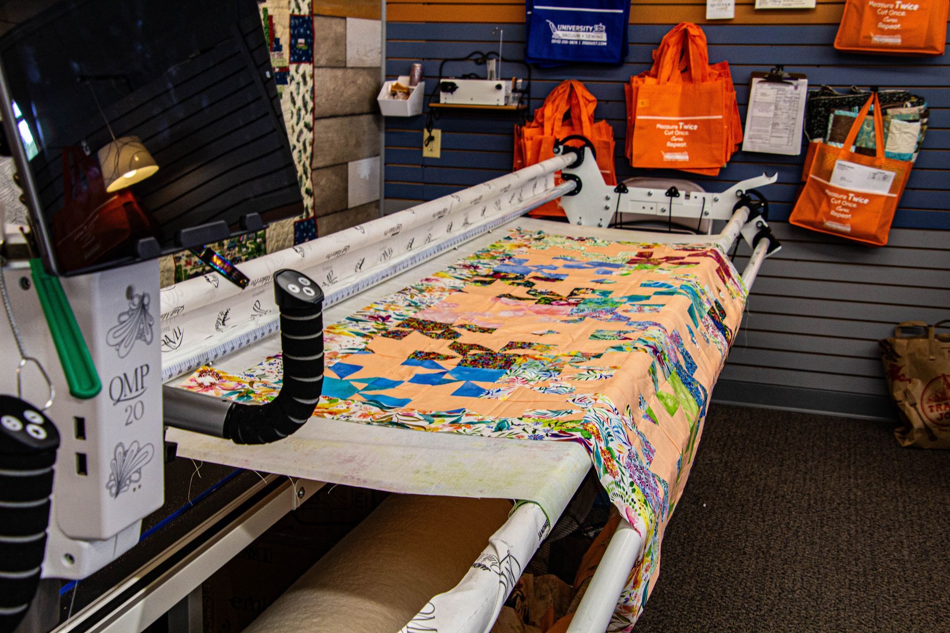 Quilting machine with fabric in a shop. Colorful quilt on the machine's frame, surrounded by orange bags and sewing equipment.