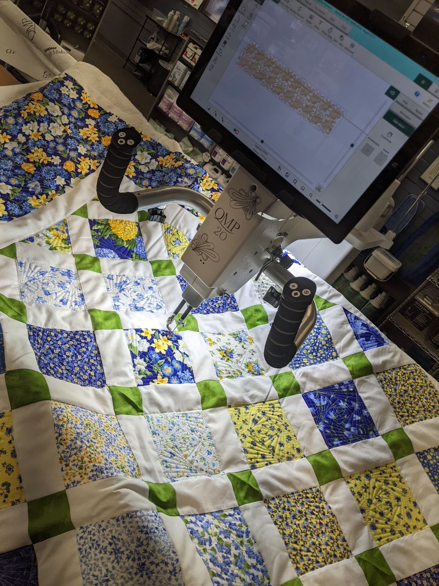 Quilt being stitched by a machine. Blue, yellow, and green patterned fabric. Computer screen visible in the background.
