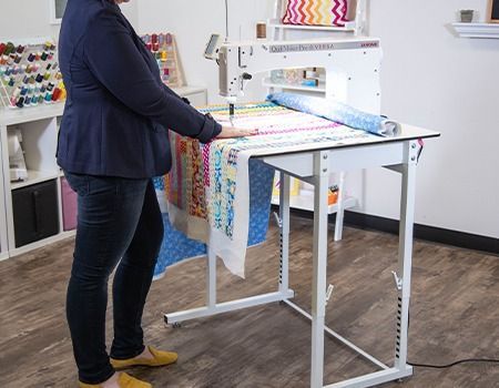 Woman quilts on a long arm machine, indoors. She wears a navy blazer, jeans, and yellow shoes.