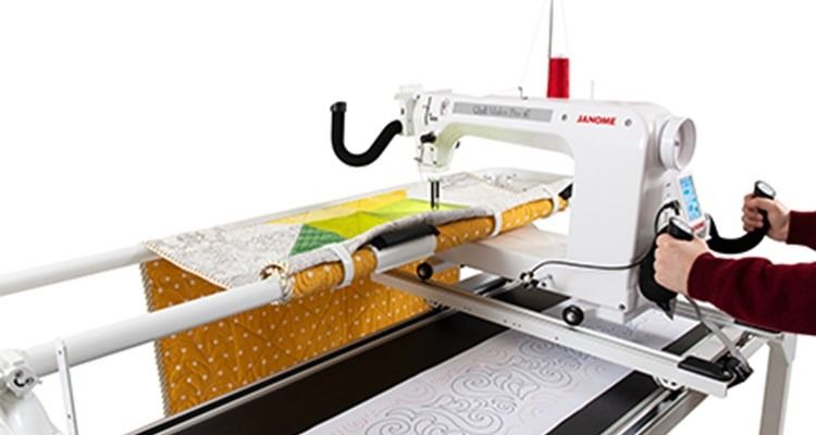 White quilting machine on a frame; person's hands operating it. Quilt in progress, with yellow and patterned fabric.