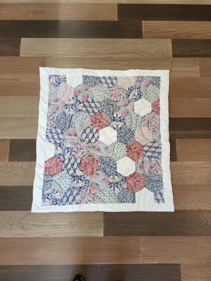 Small quilt with hexagonal patches in shades of pink, blue, and white on a wooden floor.
