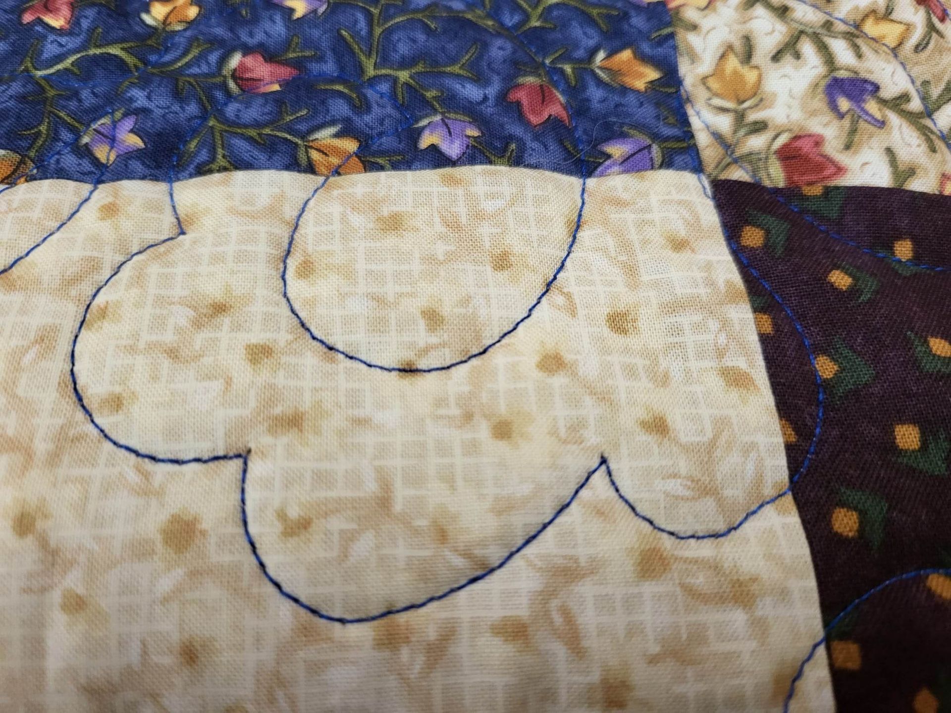 Close-up of quilted fabric. Beige, blue, and purple floral patterns with blue stitching.