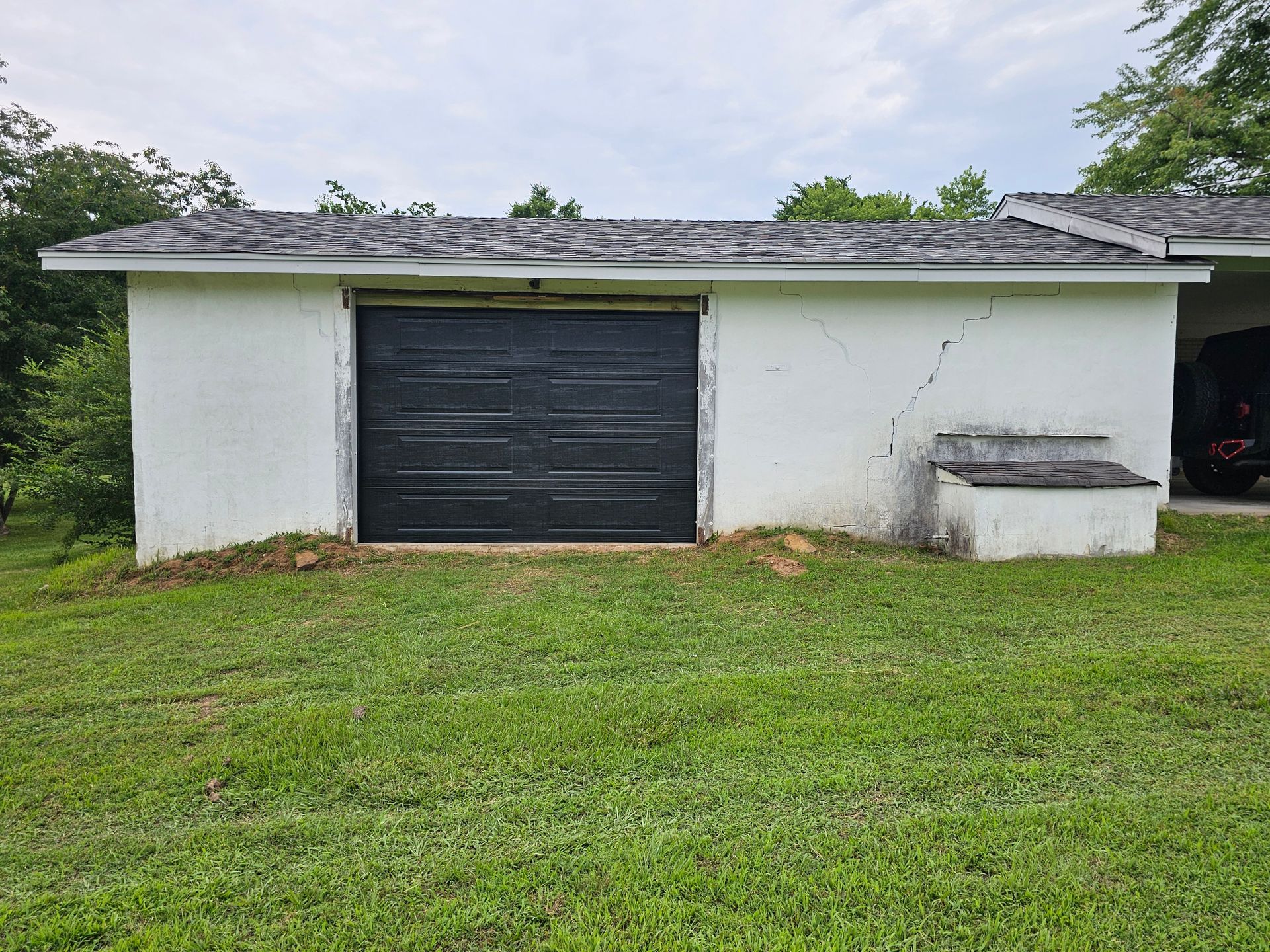 A white garage with a black garage door is sitting in the middle of a lush green field.