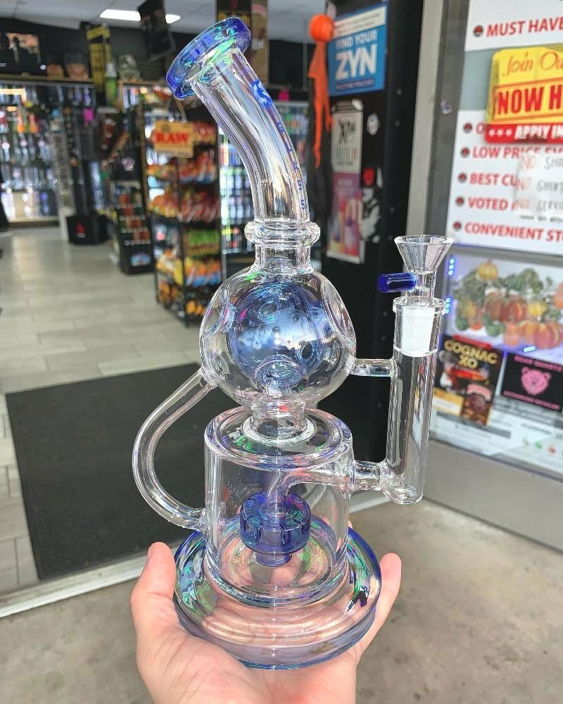 A person holding a blue and clear glass water pipe with a circular chamber, inside a store.