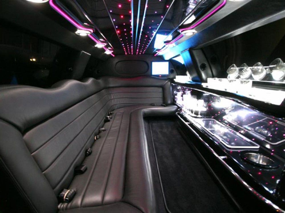 Interior of a limousine. Black leather seating, mirrored ceiling with colorful lights, and a bar area with glasses.