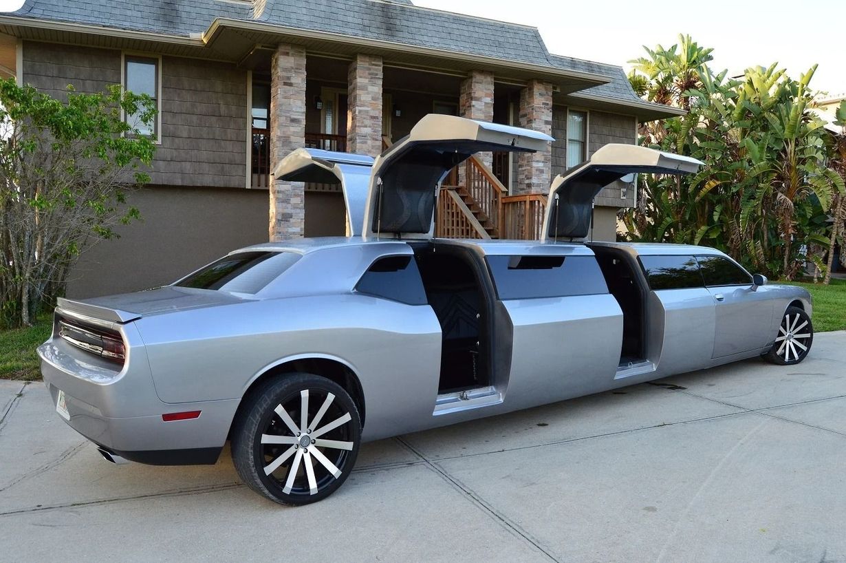 Silver Dodge Challenger limousine with gull-wing doors open, parked on a driveway in front of a house.
