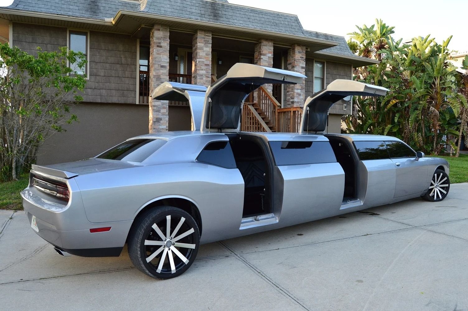 Silver Dodge Challenger limousine with gull-wing doors open, parked on a driveway in front of a house.
