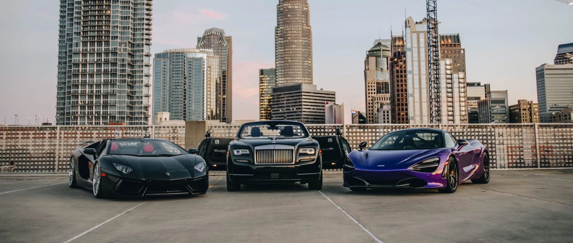 Three luxury cars parked on a rooftop overlooking a cityscape. Black Lamborghini, Rolls-Royce, and blue McLaren.