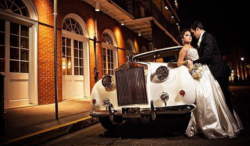 Bride and groom pose near a white vintage car at night in front of a brick building.