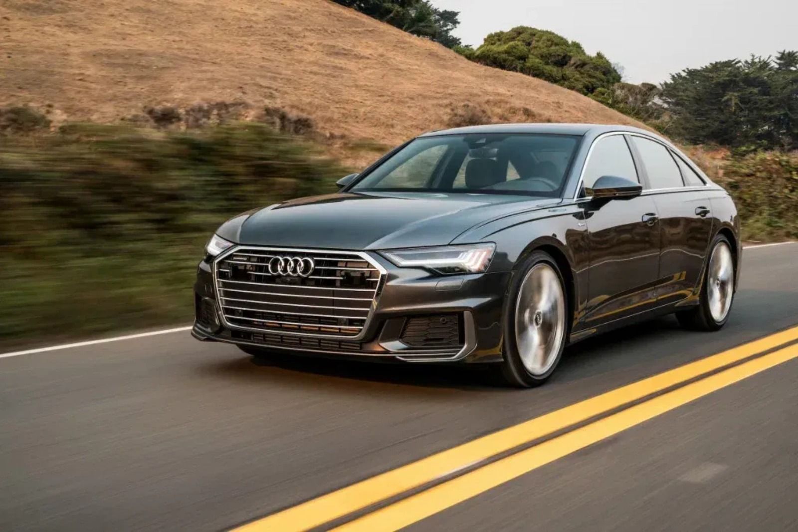 Dark gray Audi A6 sedan driving on a two-lane road with a blurred hillside in the background.