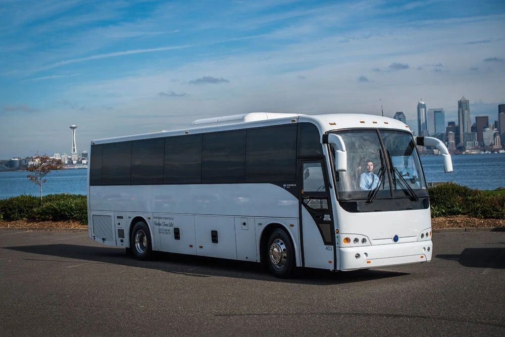 White tour bus parked on asphalt; city skyline in background.