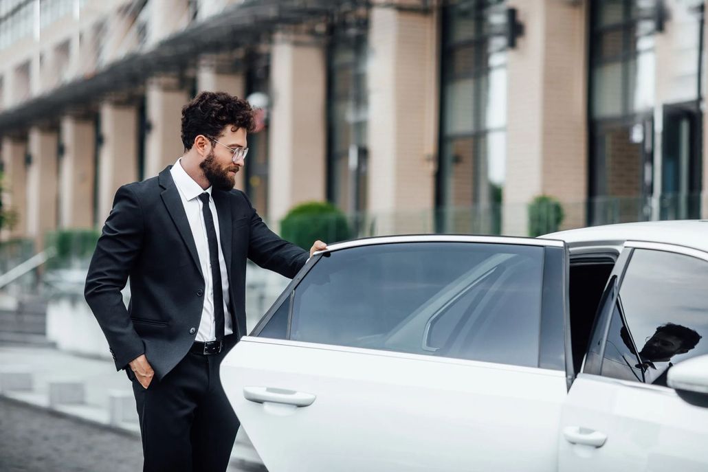 Man in suit opening car door. Building in background.
