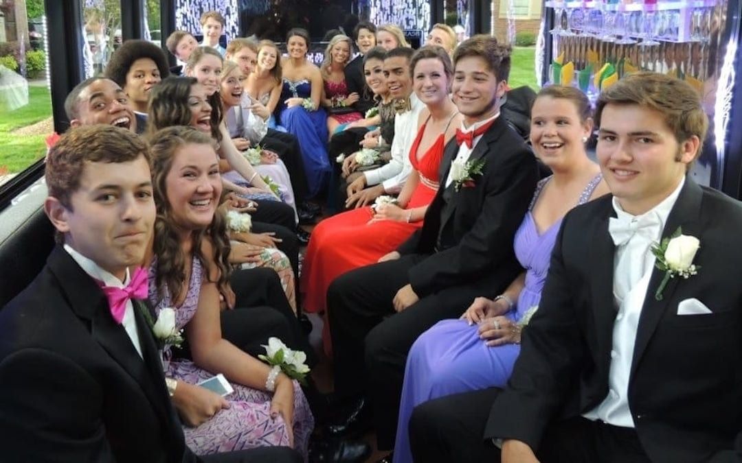 Teenagers in formal attire sit on a bus, smiling, ready for prom.