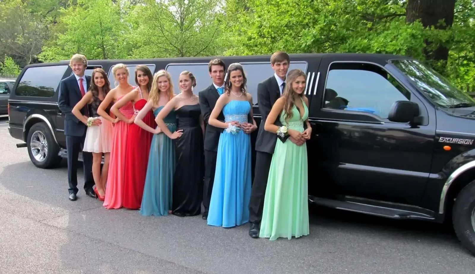 Group of teens in formal wear pose by a black limousine outdoors.