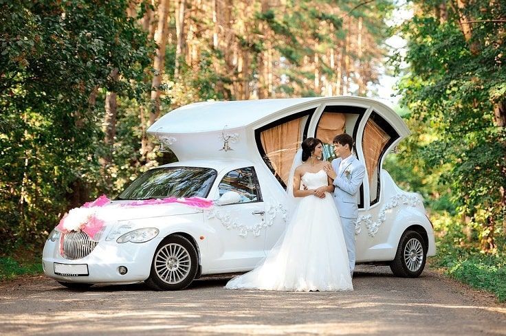 Bride and groom pose by a decorated white car on a road in a forest.