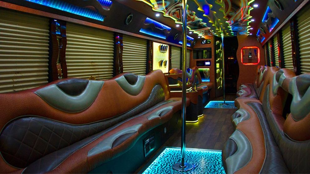 Interior of a party bus with orange, brown, and gray seating, a dance pole, and neon lights.