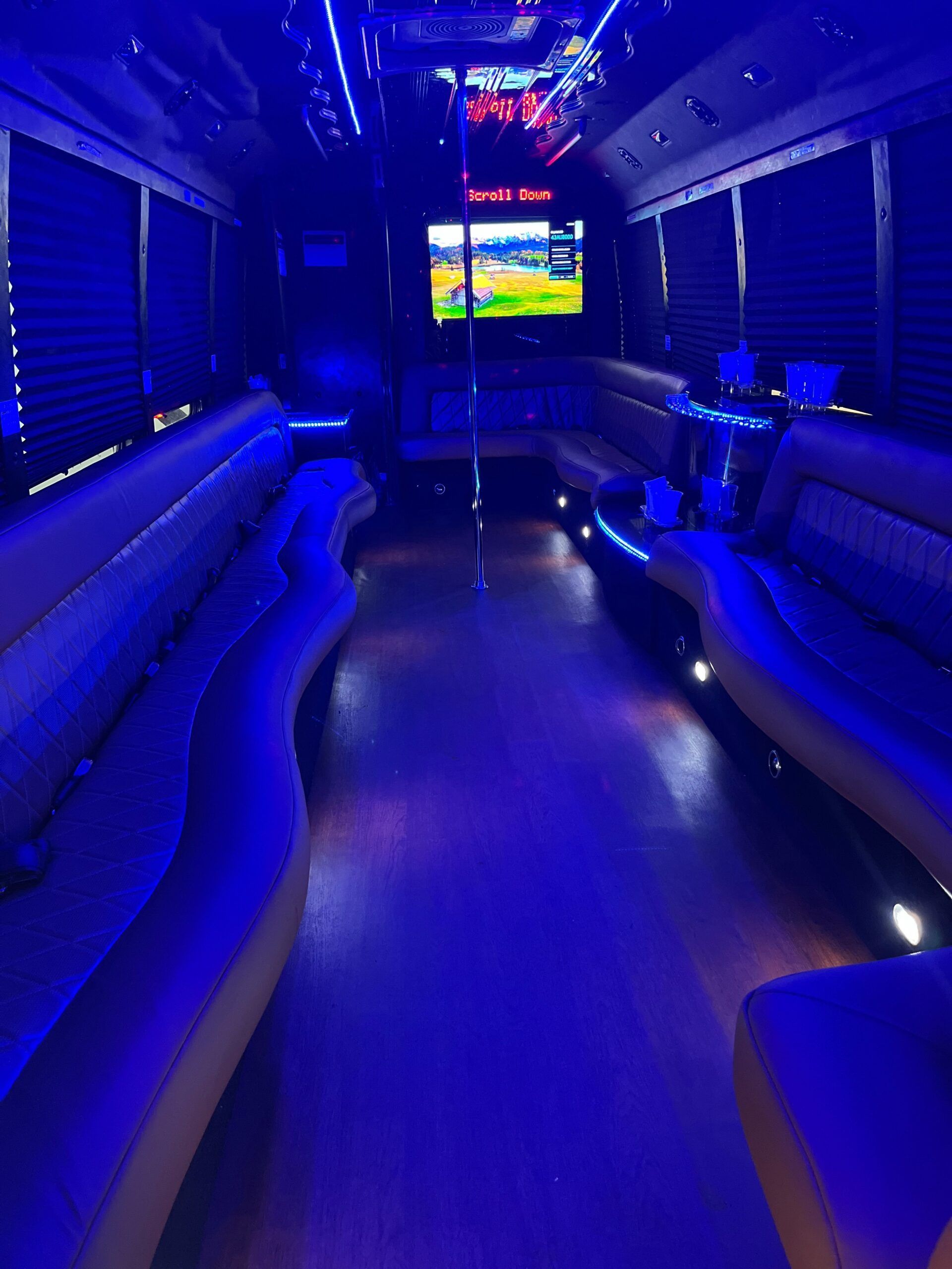 Interior of a party bus with blue lighting, seating, a pole, and a TV screen displaying a video.