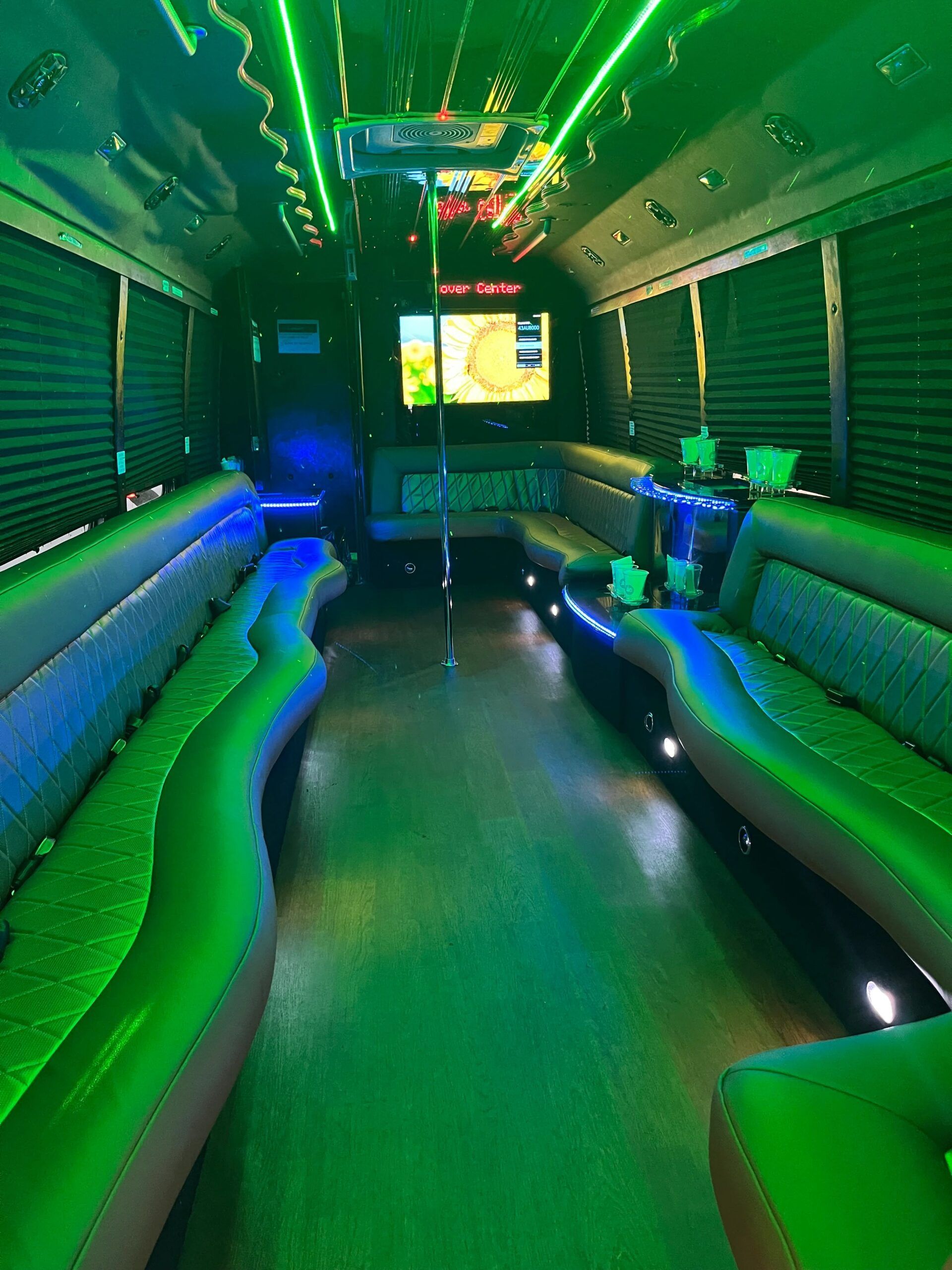 Interior of a party bus with green and blue LED lighting, seating along the sides, and a screen in the back.