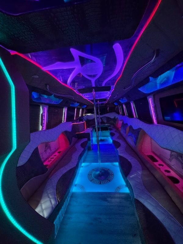Inside of a party bus with neon blue, pink, and purple lights.  Center pole and built-in seating.