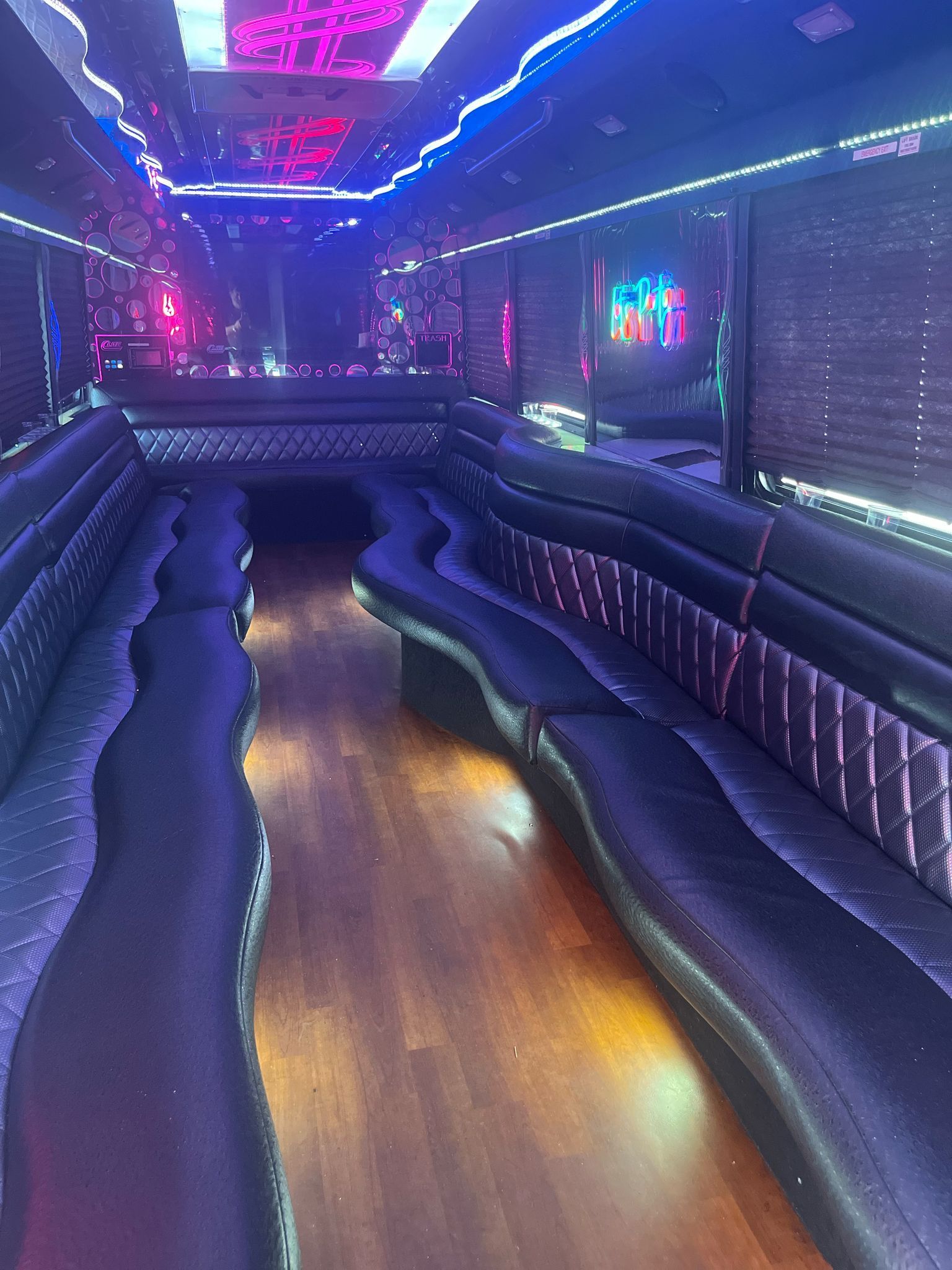 Interior of a party bus with black wraparound seating, wood floor, and neon lights.