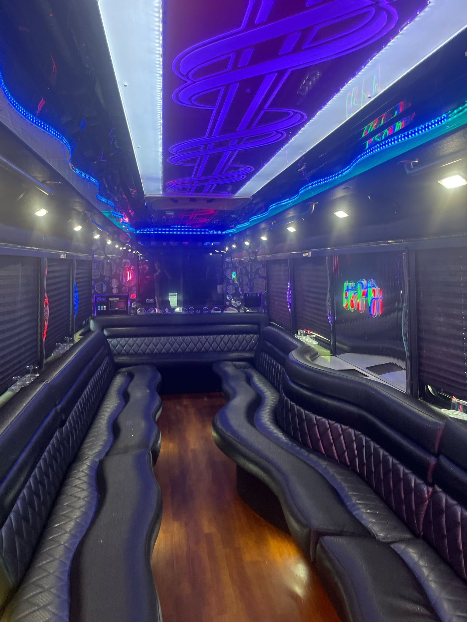 Interior of a party bus with black leather seating, wood floor, and neon lights on the ceiling.