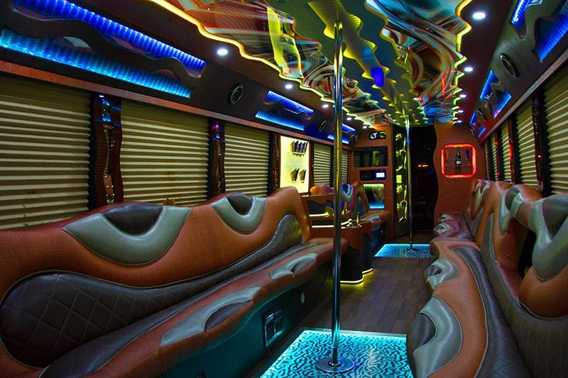 Interior of a party bus with orange and brown seating, a pole, and colorful LED lights.