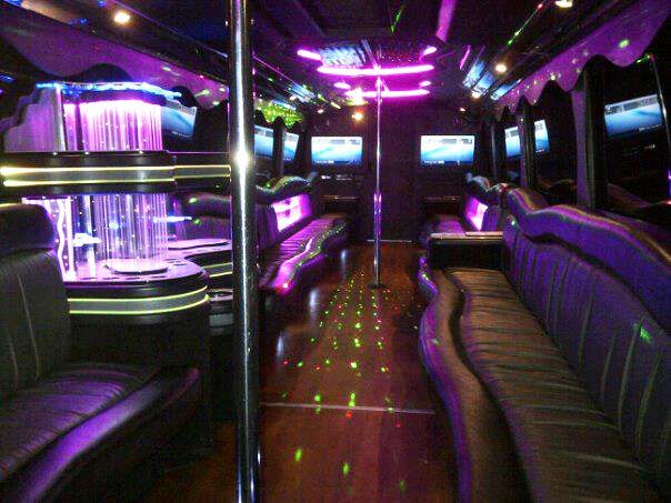 Inside a party bus: purple and blue lights, seating, a dance pole, and screens.
