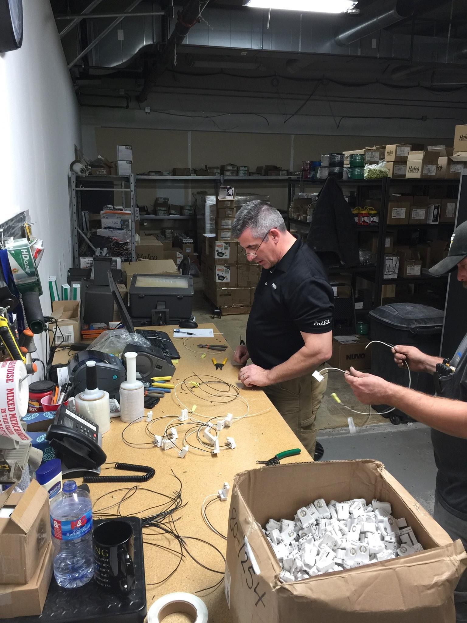 Two men assemble wiring components at a workbench in a workshop. Boxes and tools are visible.