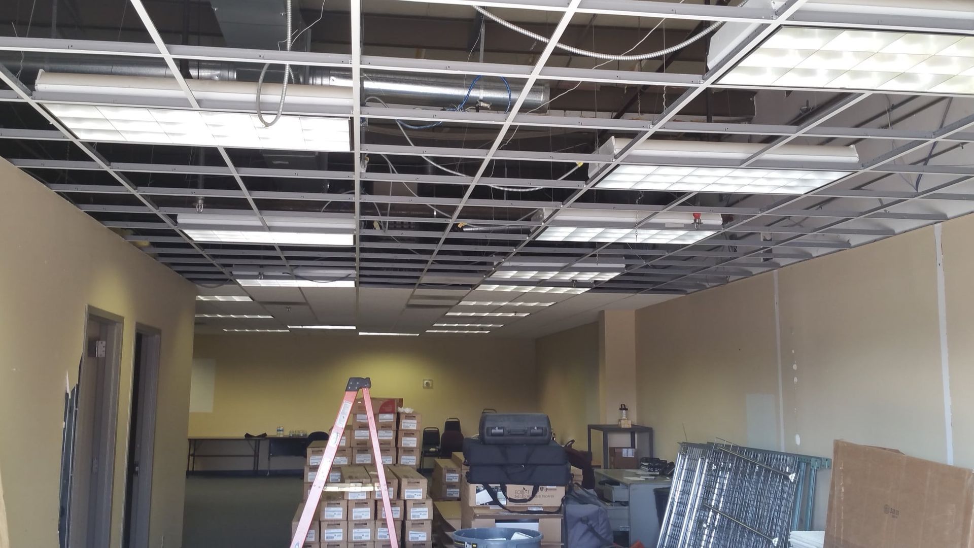 Interior view of a construction site, open ceiling with fluorescent lights, ladder, and materials.