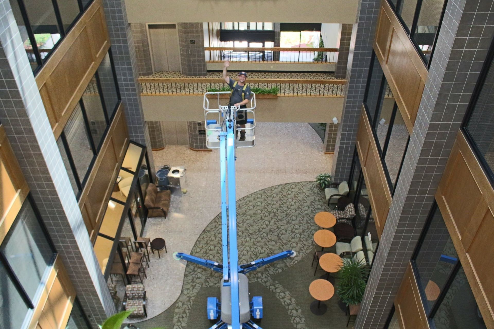 A person on a blue lift cleaning windows in a multi-story building lobby with seating and plants.