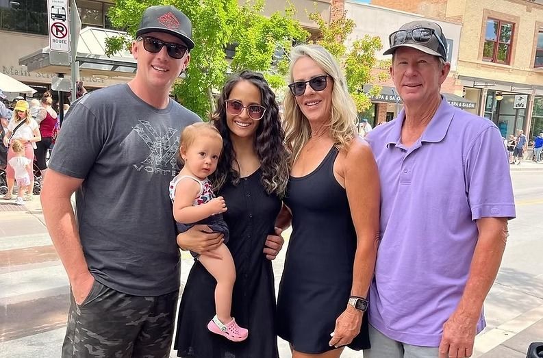 Four people and a toddler posing outdoors on a sunny street, smiling at the camera.